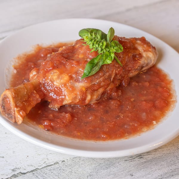 Braised lamb shank in a rich red tomato sauce, garnished with fresh basil on a white plate.