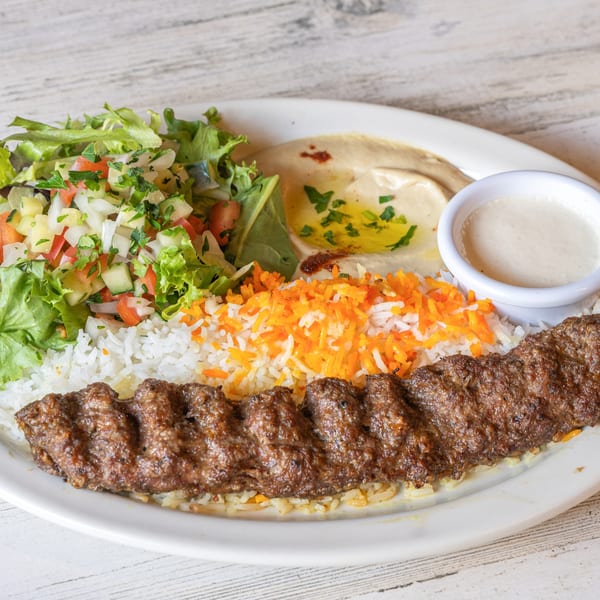 A Mediterranean plate with a long kebab, rice, salad, hummus, and a white dipping sauce.