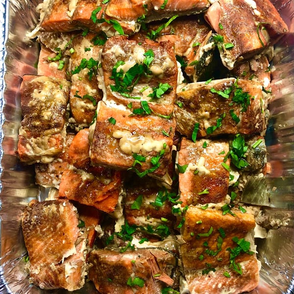 Close-up of baked salmon pieces topped with creamy sauce and fresh parsley in a foil pan.